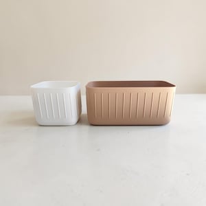 Op de afbeelding: Een kleine witte vierkante opbergbak en een grotere rechthoekige beige opbergbak. Beide bakken hebben een gladde matte afwerking en verticale ribbeldetails langs de buitenwanden, met afgeronde hoeken en een open bovenkant.