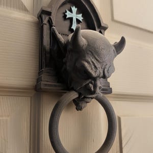 May include: A dark grey, 3D-printed door knocker featuring a sculpted gargoyle head with horns and pointed ears. The gargoyle is mounted on a gothic-style arched backplate with a light blue cross emblem. A circular ring hangs from the gargoyle's mouth.