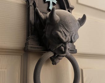 May include: A dark gray, 3D-printed door knocker featuring a sculpted gargoyle head with horns and pointed ears. The gargoyle is mounted on a gothic-style arched backplate with a light blue cross emblem. A circular ring hangs from the gargoyle's mouth.