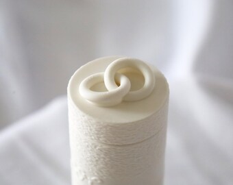 May include: A cylindrical white container with a smooth, flat top surface featuring two interlocking white rings. The body of the container has a subtle, fine-textured finish that wraps around the exterior, creating a clean and uniform appearance.