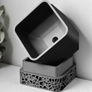 May include: A black and grey geometric planter with a square opening. The planter is made of a textured material and has a unique design.