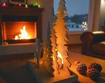 May include: A white wooden decorative piece featuring two stylized evergreen trees and a small deer silhouette mounted on a rectangular base. The design includes a hollow center space designed to hold a small tea light candle, casting a warm glow.