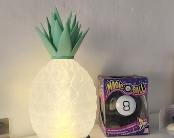 May include: A pineapple-shaped lamp with a white textured body, a light green top, and a black base, illuminated from within. Next to it sits a Magic 8 Ball toy in its original purple and black packaging with the text Magic 8 Ball visible.