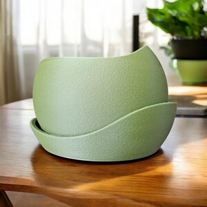 Könnte beinhalten: Ein hellgrüner, strukturierter Keramik-Blumentopf mit passender Untertasse. Der runde Topf hat ein einzigartiges geschwungenes Design am Rand. Der Topf steht auf einem Holztisch. Im Hintergrund ist eine grüne Pflanze zu sehen.