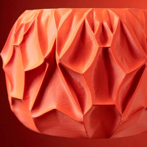 Könnte beinhalten: Nahaufnahme einer orangefarbenen dekorativen Schale mit einer geometrischen, strukturierten Oberfläche. Das Design der Schale weist rautenförmige Facetten auf, die ein Spiel von Licht und Schatten erzeugen. Die Farbe ist ein leuchtendes, warmes Orange.