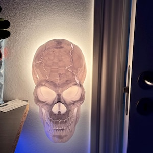 Może przedstawiać: Biała ceramiczna lampka nocna w kształcie czaszki z błyszczącymi oczami. Czaszka jest zamontowana na ścianie i jest oświetlona od tyłu.