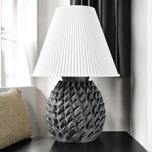 Può includere: Una moderna lampada da tavolo con paralume bianco plissettato e base grigio scuro strutturata. La base presenta un motivo geometrico a forma di diamante. La lampada è su una superficie nera, con una finestra e una tenda sullo sfondo.