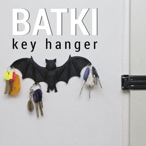 May include: A black bat-shaped key hanger mounted on a white wall, with the word "BATKI" above and "key hanger" below. Several keys hang from the bat's wings. The bat is made of a textured material.