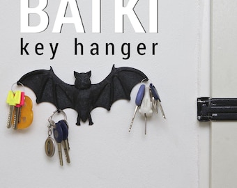 Op de afbeelding: Een zwarte vleermuisvormige sleutelhouder aan een witte muur, met het woord "BATKI" erboven en "key hanger" eronder. Verschillende sleutels hangen aan de vleugels van de vleermuis. De vleermuis is gemaakt van een textuurrijk materiaal.