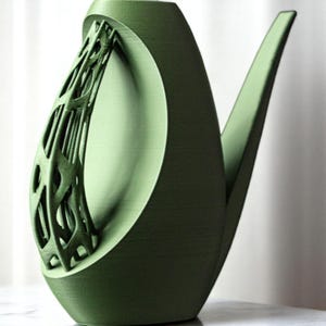Op de afbeelding: Een groene theepot van keramiek met een uniek, ingewikkeld ontwerp op de handgreep. De theepot heeft een gebogen tuit en een brede basis.