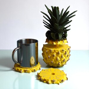 Può includere: Una tazza in acciaio inossidabile poggia su un sottobicchiere giallo con un design ad ananas. È presente anche un ananas decorativo con la parte superiore verde e il corpo giallo, insieme a un sottobicchiere separato. Gli oggetti sono su una superficie chiara.
