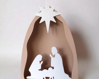 May include: A layered nativity scene decoration featuring a beige arched frame with a white glittery star at the top. Inside the arch, white silhouettes of two figures kneeling beside a small central figure are positioned against the beige background.