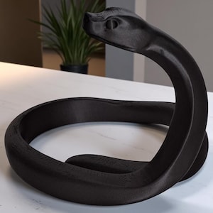 Può includere: Scultura a forma di serpente nero arrotolato. La scultura presenta una texture dettagliata e la testa sollevata. È realizzata in un materiale scuro e poggia su una superficie bianca. Il design è moderno e artistico.