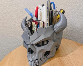 May include: A gray, 3D-printed desk organizer shaped like a horned skull with a faceted, geometric surface. The hollow interior holds various items including pens, a flashlight, a wallet, and a ruler with visible text and markings.