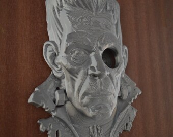May include: A gray, three-dimensional wall-mounted sculpture depicting a stylized human face with a prominent forehead, textured hair, and a hollowed-out left eye socket. The figure features a high collar and visible neck bolts on both sides.