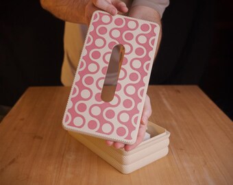 May include: A rectangular tissue box cover featuring a repeating pattern of pink circles connected by white curved lines. The cover has a central elongated oval opening and a textured, cream-colored base section visible underneath.