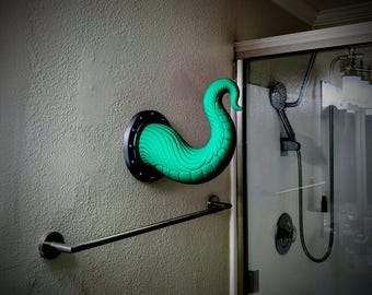 May include: A bright green, curved tentacle-shaped wall hook mounted on a circular black base with visible screw holes. The tentacle features a segmented, ridged texture and extends outward from the wall, positioned above a metal towel bar.
