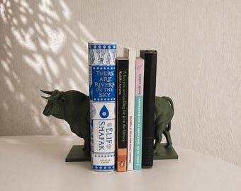 May include: A set of two dark green bull-shaped bookends holding a row of five books. The books include titles such as There Are Rivers in the Sky by Elif Shafak, What You Are Looking For Is in the Library, and I Want to Die but I Want to Eat Tteokbokki.