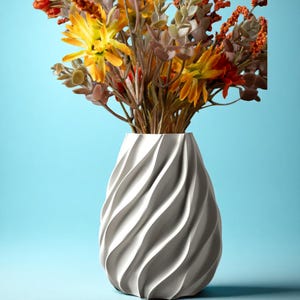 Può includere: Un vaso bianco strutturato con un motivo a spirale contiene un bouquet di fiori artificiali colorati. I fiori presentano tonalità gialle, arancioni e marroni. Il vaso è posto su uno sfondo azzurro chiaro, creando un contrasto visivo.
