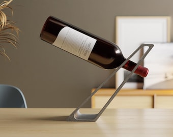May include: A dark gray, angular, open-frame wine bottle holder made of a textured material. A wine bottle with a white label reading Fiera Vinea is balanced at an angle within the frame, with the neck pointing downward toward the right.