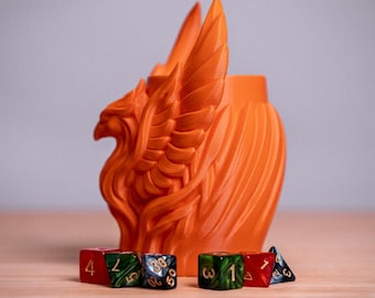 May include: A bright orange 3D-printed dice cup featuring a sculpted phoenix with raised wings on the exterior. The cup is surrounded by a collection of multi-sided gaming dice in red, green, blue, and black with gold-colored numbers.