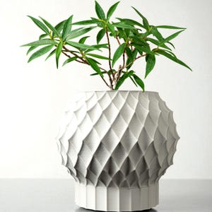 Op de afbeelding: Een witte, getextureerde, bolvormige vaas met een geometrisch ruitpatroon. De vaas bevat een groene plant met lange, slanke bladeren. De vaas heeft een brede basis en een afgeronde bovenkant, wat een moderne uitstraling creëert.