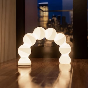 Puede incluir: Una lámpara moderna blanca en forma de arco con una serie de luminarias esféricas conectadas. La lámpara está iluminada, proyectando un brillo cálido sobre el suelo de madera. El fondo muestra un paisaje urbano al anochecer.