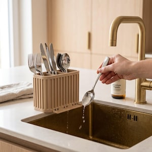 May include: A beige, rectangular, ribbed plastic cutlery drainer with multiple compartments holding stainless steel forks, knives, and spoons. The drainer sits on a white countertop next to a brass kitchen faucet and a brass sink basin.
