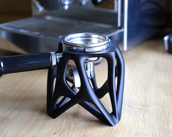 May include: A black espresso portafilter stand with a matte finish and a geometric, open-frame structure. The stand supports a stainless steel portafilter basket with a black handle, positioned horizontally for stability during coffee preparation.