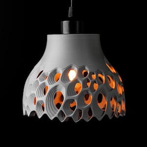 Op de afbeelding: Een grijze, moderne hanglamp met een uniek, opengewerkt ontwerp. De lampenkap heeft een reeks cirkelvormige uitsparingen die een warme, oranje gloed onthullen van de lichtbron binnenin. De armatuur hangt aan een zwart snoer tegen een zwarte achtergrond.