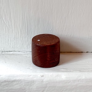 May include: A cylindrical wooden knob with a smooth, dark brown finish and a visible wood grain pattern. The top surface of the knob features a single, small, circular white inlay positioned near the edge, providing a distinct contrast.