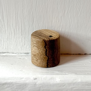 May include: A cylindrical wooden block featuring a natural light brown grain pattern with a distinct dark vertical crack running down the side and a small circular hole visible on the flat top surface, showcasing the raw texture of the wood.
