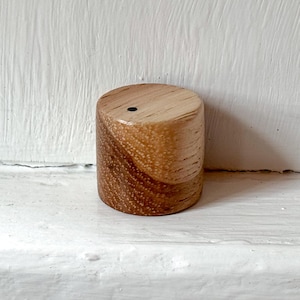 May include: A cylindrical wooden knob featuring a natural light and medium brown wood grain pattern with a smooth, polished finish. The top surface of the knob displays a single, small, centered black circular dot detail on the wood grain.