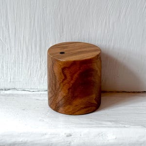 May include: A cylindrical wooden shaker featuring a smooth, polished finish with visible natural grain patterns in shades of medium and dark brown. The top surface is flat and circular, displaying a single small, centered black circular hole.