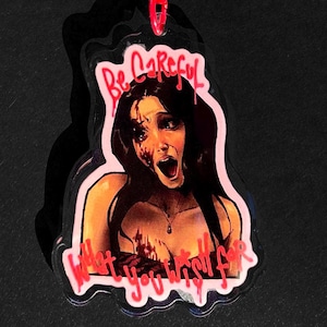 May include: A clear acrylic keyring featuring a graphic illustration of a person with long dark hair, a wide-eyed expression, and blood-like markings on their face and chest. The text "Be Careful" and "What you wish for" is written in red.