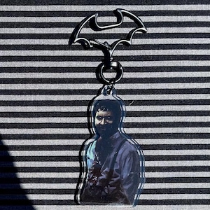 Puede incluir: Un llavero con temática de Batman con un símbolo de murciélago plateado y un colgante de acrílico transparente con la imagen de una persona. El colgante tiene una imagen detallada de una persona con una camisa azul.
