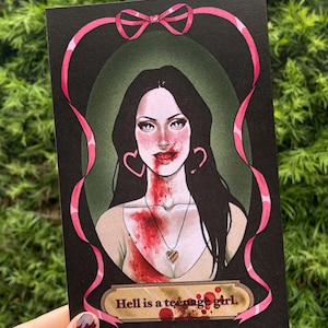May include: A black and white illustration of a woman with long dark hair. She is wearing a white tank top and a gold heart necklace. She has blood on her face and chest. The background is a dark green with a pink ribbon around the frame. The text "Hell is a teenage girl." is at the bottom of the image.