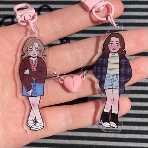 Puede incluir: Dos llaveros acrílicos transparentes con ilustraciones de dibujos animados de mujeres jóvenes. Un llavero muestra a una mujer con una chaqueta roja y una falda vaquera, el otro con una camisa a cuadros y pantalones cortos. Un dije de corazón rosa conecta los llaveros.