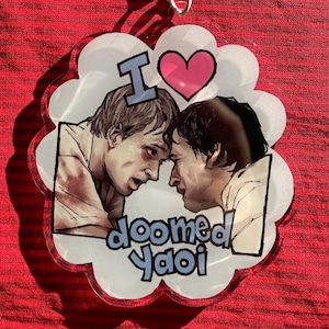 Könnte beinhalten: Ein klarer, wolkenförmiger Schlüsselanhänger mit einer Grafik von zwei Männern, die sich gegenüberstehen, mit den Worten "I" und einem rosa Herzen darüber. Darunter steht der Text "doomed Yaoi". Der Hintergrund ist rot.