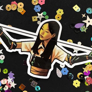 May include: A sticker featuring a stylized illustration of a woman in a white shirt and black gloves, holding a white string. The sticker is surrounded by colorful sequins and confetti on a black background.