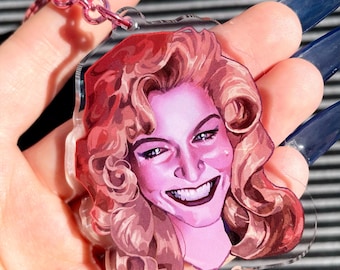 May include: A clear acrylic keyring featuring a smiling woman with pink skin and curly auburn hair. The keyring has a pink metal chain and ring. The woman's portrait is a vibrant illustration with a detailed face and dark lipstick.