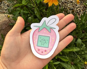 May include: A pink and green strawberry-shaped sticker with a cartoon strawberry character on a screen. The sticker is held in a hand.