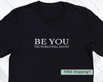 May include: Black crew neck t-shirt with the white text "BE YOU" above "THE WORLD WILL ADJUST". The shirt is a simple design, perfect for casual wear.