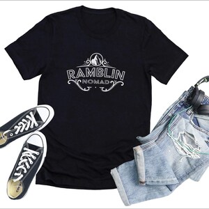 Puede incluir: Camiseta negra con un dise&ntilde;o gr&aacute;fico blanco que dice "Ramblin Nomad" con un pico de monta&ntilde;a y un dise&ntilde;o floral. La camiseta est&aacute; colocada sobre una superficie blanca con un par de zapatillas negras y unos vaqueros azules con unos auriculares verdes.