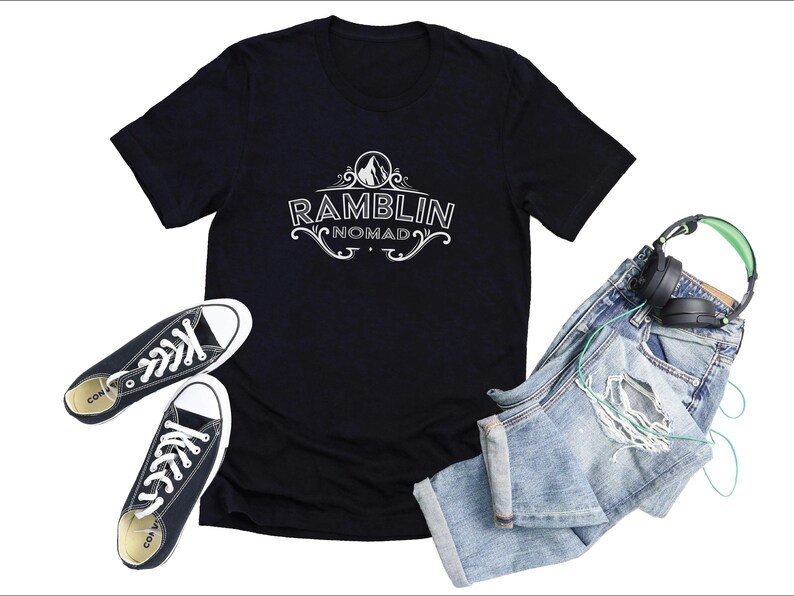 Puede incluir: Camiseta negra con un dise&ntilde;o gr&aacute;fico blanco que dice "Ramblin Nomad" con un pico de monta&ntilde;a y un dise&ntilde;o floral. La camiseta est&aacute; colocada sobre una superficie blanca con un par de zapatillas negras y unos vaqueros azules con unos auriculares verdes.