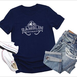 Puede incluir: Una camiseta azul marino con un dise&ntilde;o gr&aacute;fico blanco que dice "Ramblin Nomad" en una fuente de estilo vintage. La camiseta se combina con un par de jeans de mezclilla azul claro y zapatillas blancas.