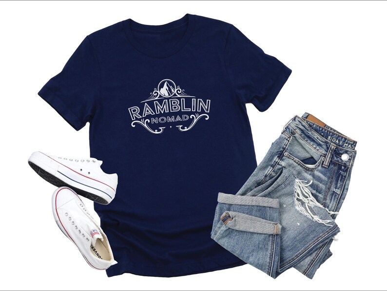 Puede incluir: Una camiseta azul marino con un dise&ntilde;o gr&aacute;fico blanco que dice "Ramblin Nomad" en una fuente de estilo vintage. La camiseta se combina con un par de jeans de mezclilla azul claro y zapatillas blancas.