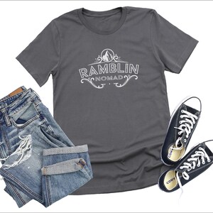Puede incluir: Una camiseta gris antracita con un dise&ntilde;o gr&aacute;fico blanco que dice "Ramblin Nomad" con una monta&ntilde;a y un dise&ntilde;o floral.