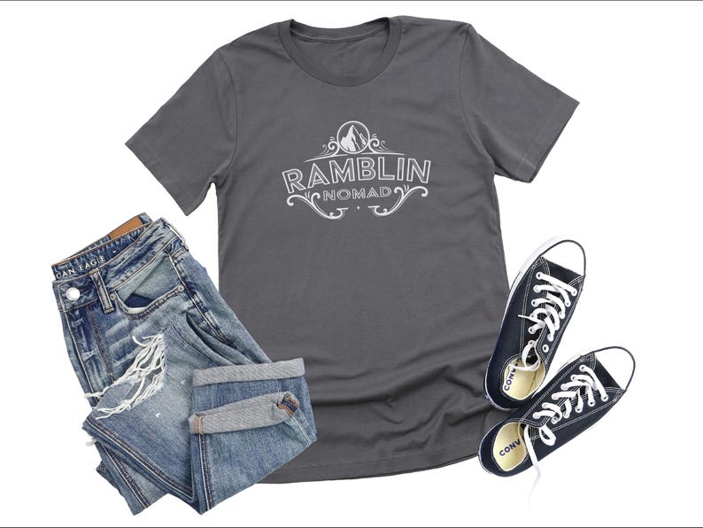 Puede incluir: Una camiseta gris antracita con un dise&ntilde;o gr&aacute;fico blanco que dice "Ramblin Nomad" con una monta&ntilde;a y un dise&ntilde;o floral.
