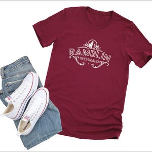 Puede incluir: Una camiseta color granate con texto blanco que dice "Ramblin Nomad". El texto est&aacute; en una fuente decorativa e incluye un peque&ntilde;o gr&aacute;fico de un pico de monta&ntilde;a.